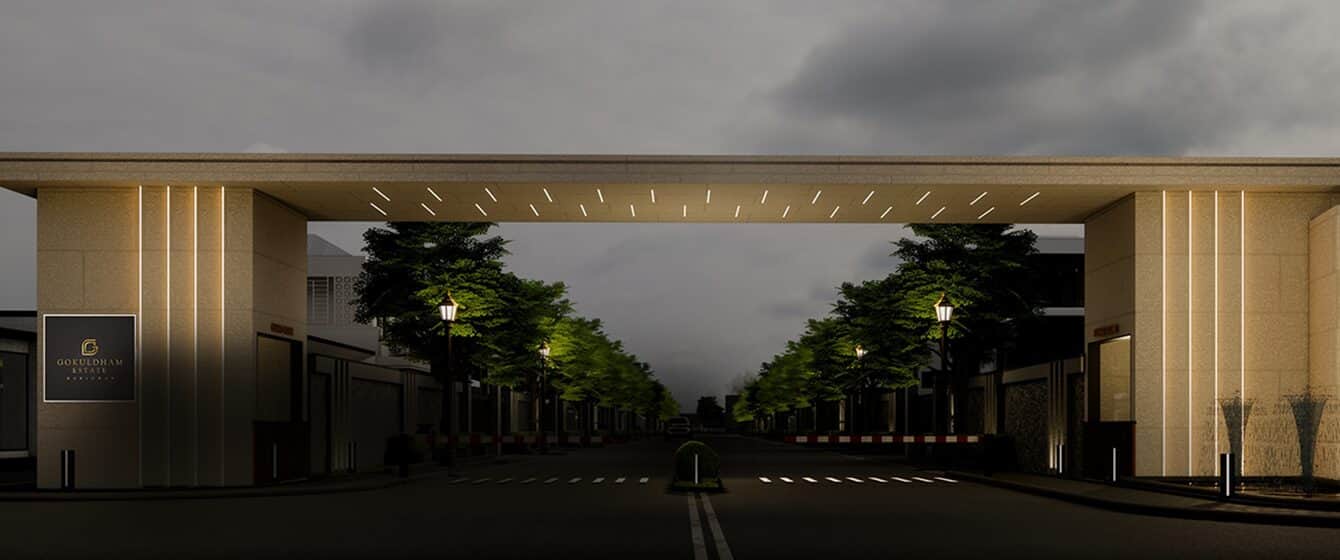 Gokuldham Estate entrance gate in Haridwar at dusk, illuminated stone piers with a lit driveway receding into planted avenues.